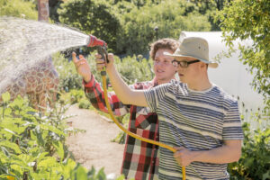 two men outside. One is watering plants with a hose.