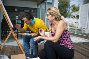 A picture of a young man and a women. The women is assisting while the man is painting a canvas.